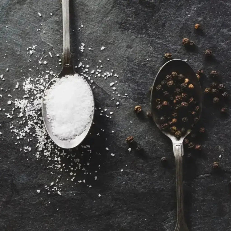 Salt and pepper on two different spoons against a black background.